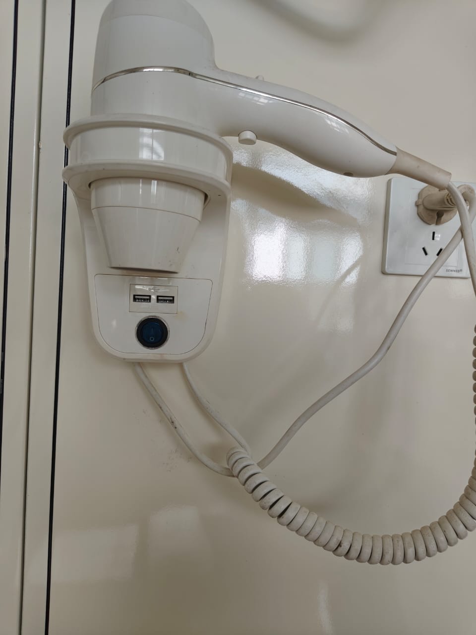 Plug-in wall-mounted hotel hair dryer base showing the mains cable opening and fixed coiled cord connection